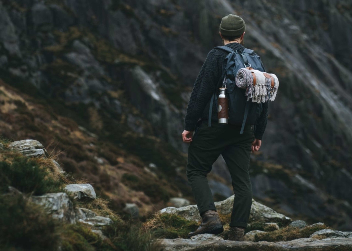 A man is walking up a mountain in Wales with a backpack on that carries a recycled wool camper blanket and adventure bottle for gym