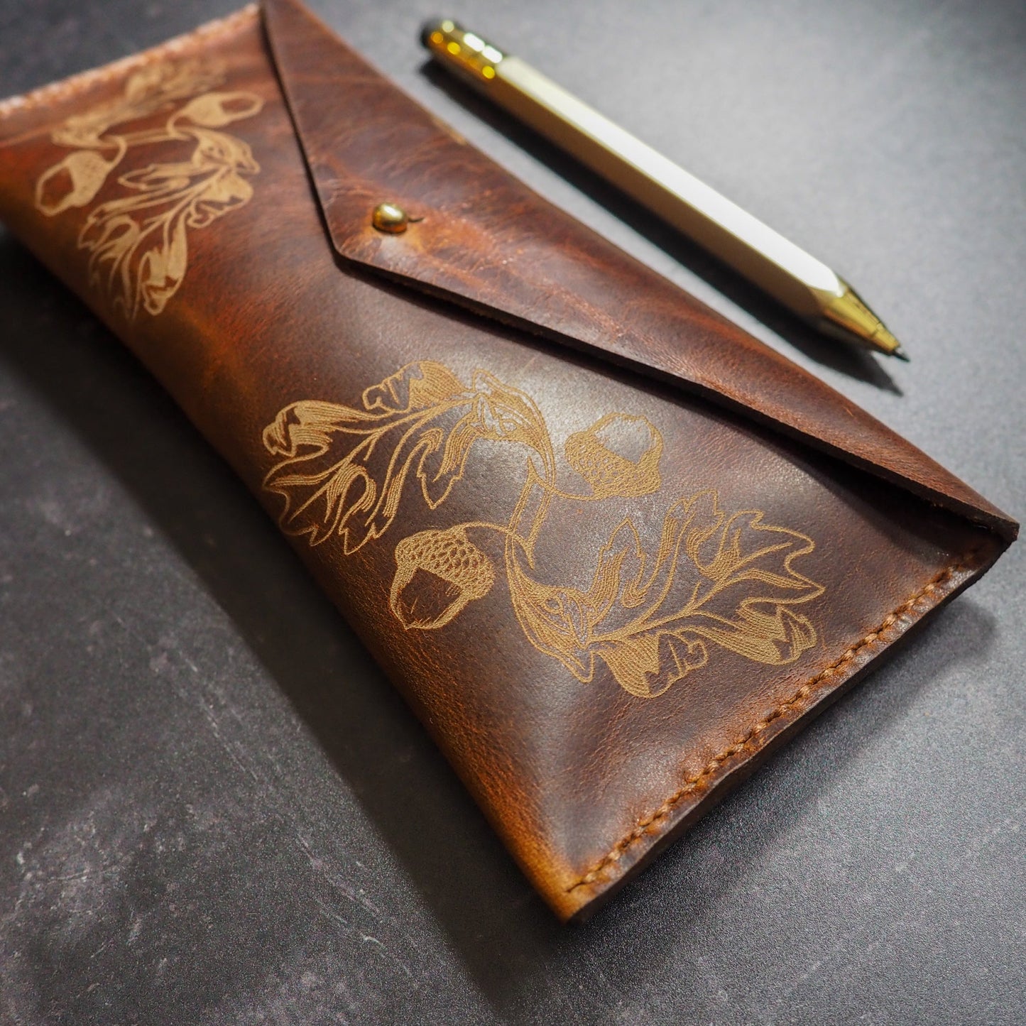 Brown leather pouch with oak leaf embossed design on a gray surface