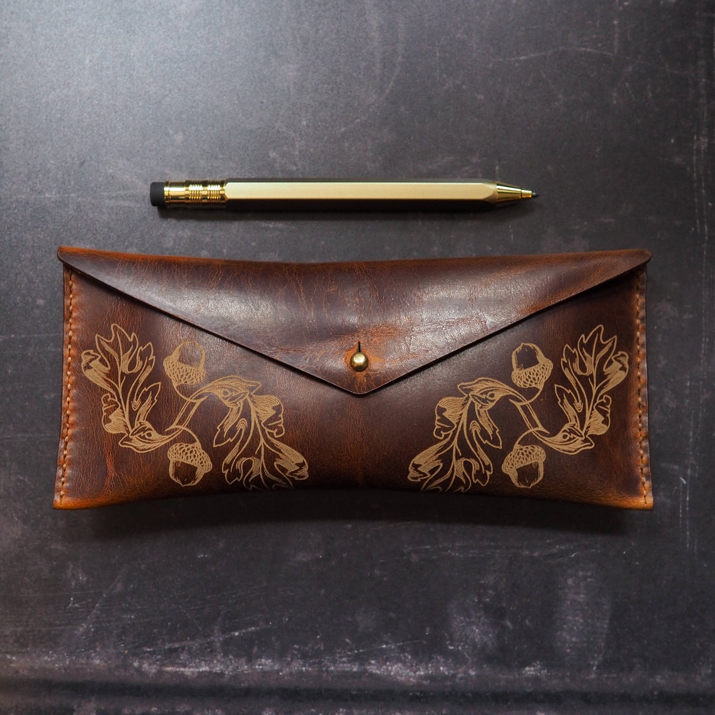 Brown leather pouch with oak leaf embossing and gold pen on a dark textured surface