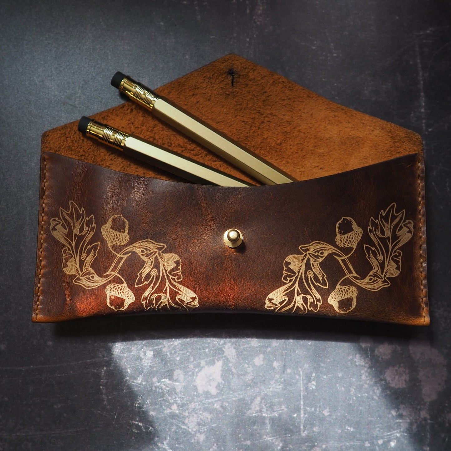 Brown leather pouch with floral patterns containing two pens on a dark textured surface