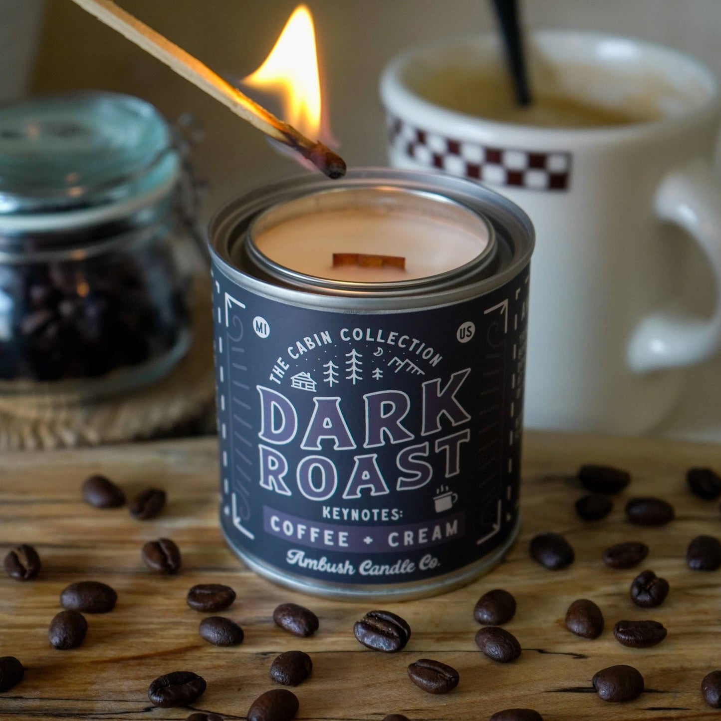 An 8oz soy candle with a label reading 'DARK ROAST COFFEE + CREAM' is lit, placed on a wooden surface with coffee beans scattered around. In the background, there is a cup of coffee and a candle is visible in the top right corner.