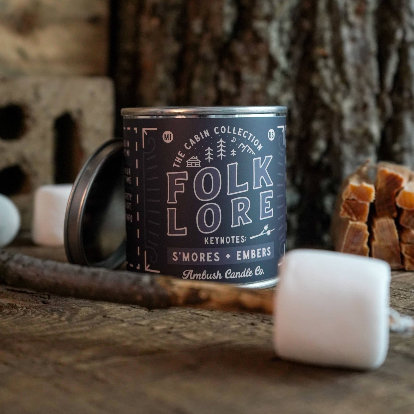 A can of 'Folklore S'mores + Embers' soy candle by Ambush Candle Co. is displayed with marshmallows and embers in the background, suggesting the scent profile of the candle.