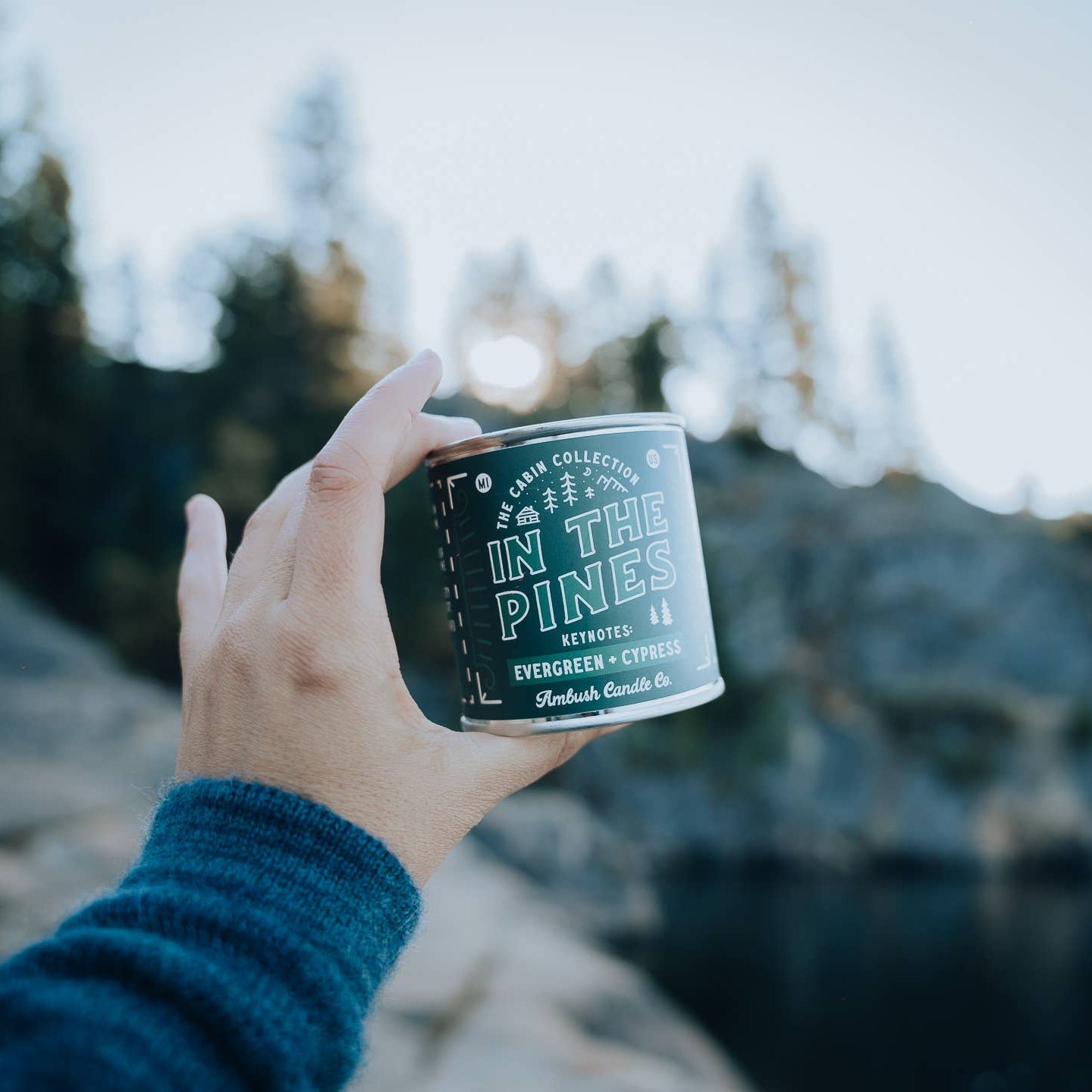 A hand holding an 8oz soy candle with 'In The Pines' and other branding on the label, in a scenic outdoor setting with trees and a body of water in the background.