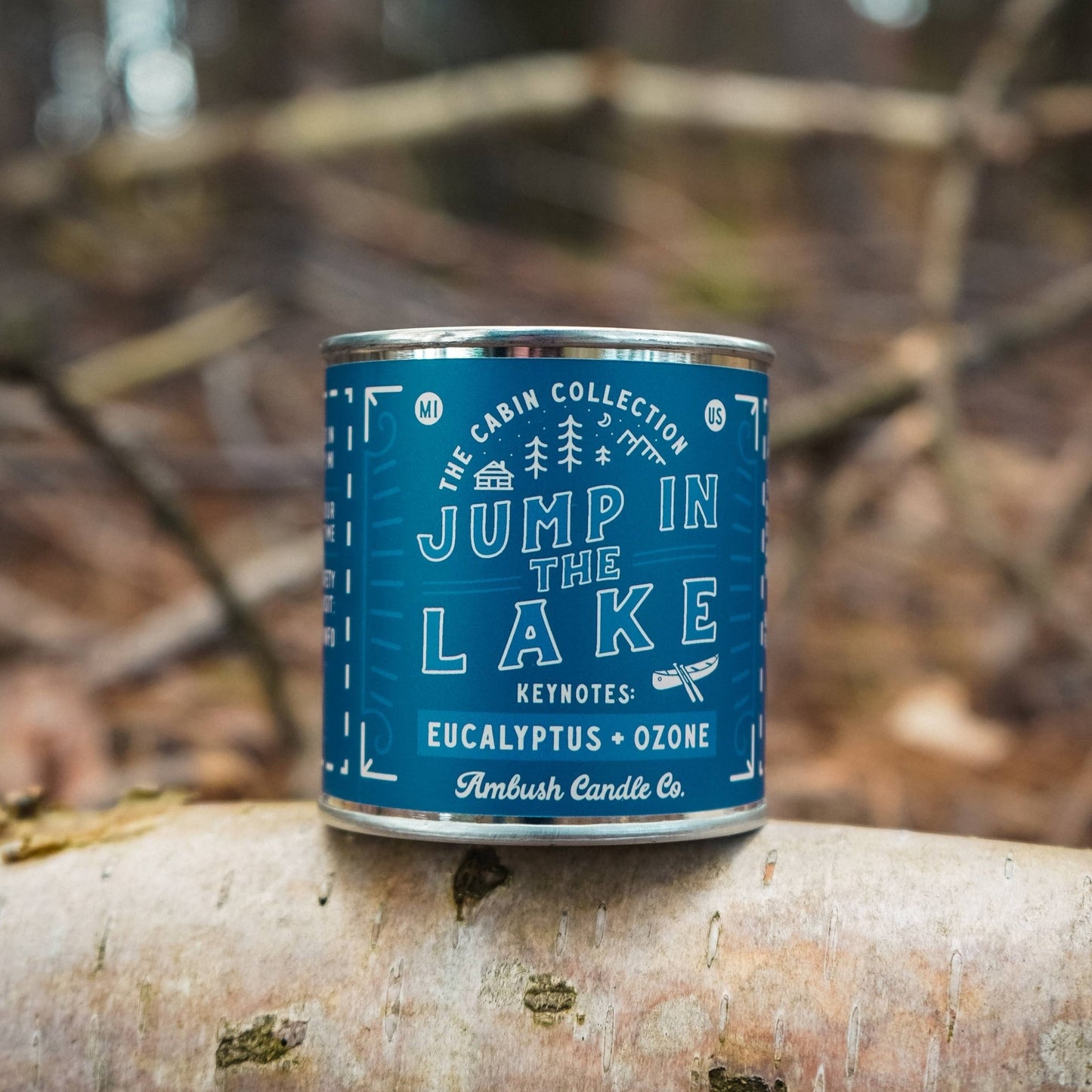 A blue metal candle tin labeled 'Jump In The Lake' with 'Eucalyptus + Ozone' scent, placed on a wooden surface in a natural outdoor setting.