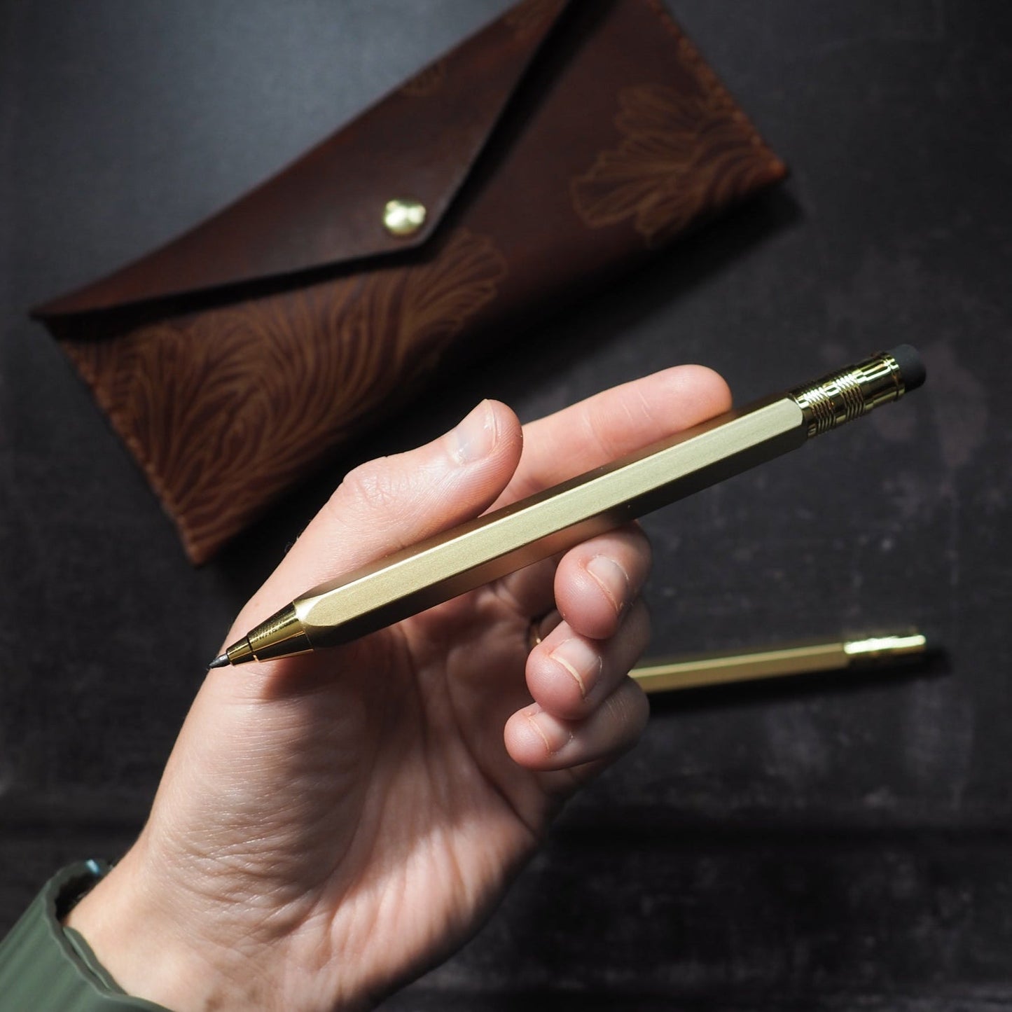 Hand holding a gold pen with a brown leather case on a dark background