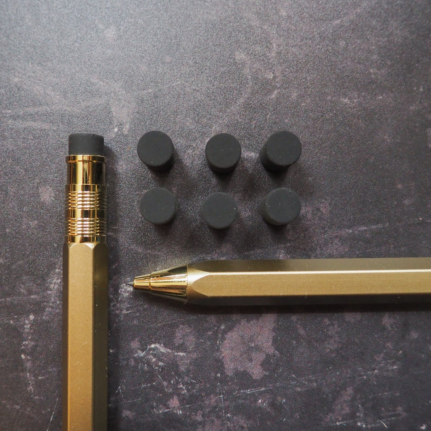 Gold pen and mechanical pencil with black erasers on a dark surface