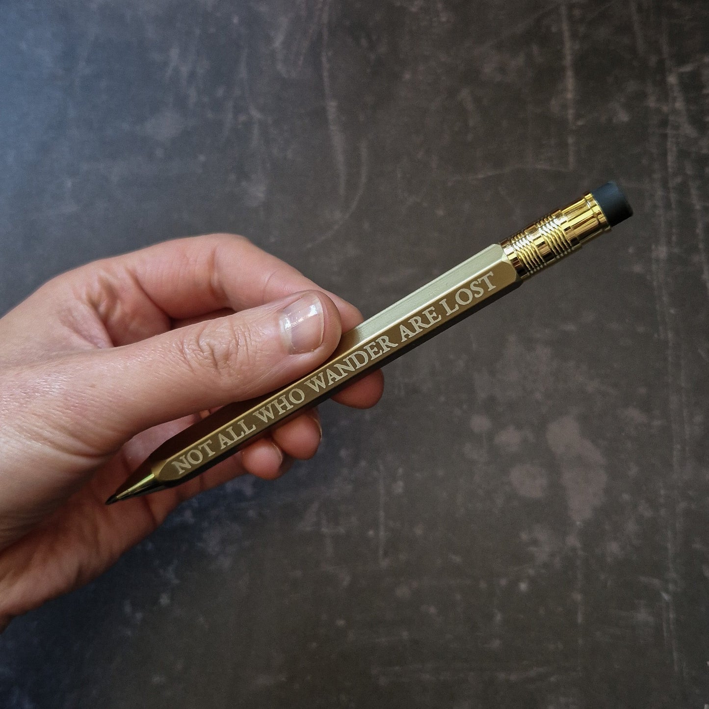 Hand holding a gold pencil with engraved text on a dark textured background