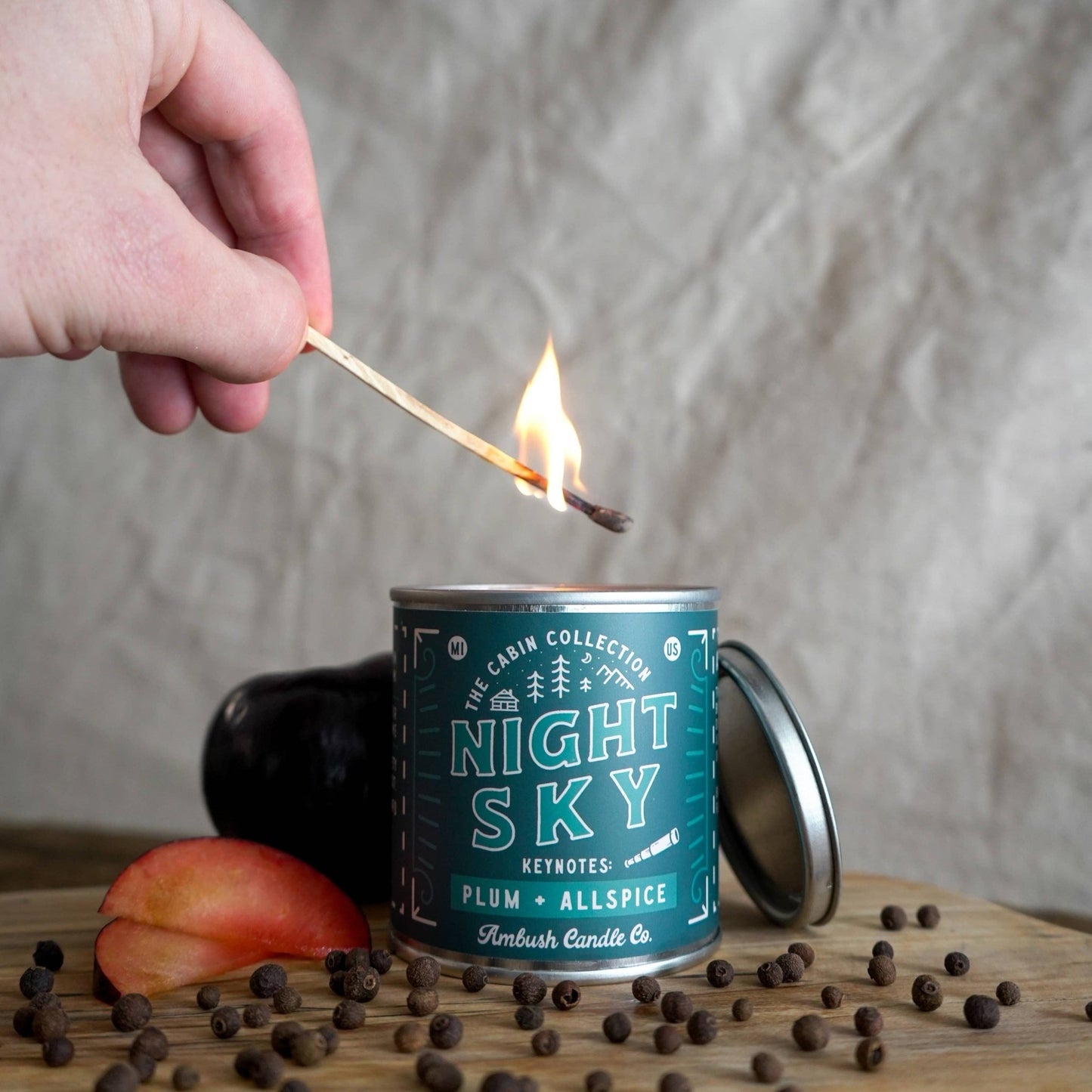 A hand is holding a lit soy candle with plum and allspice fragrance, placed on a wooden surface with a label that reads 'NIGHT SKY Plum & Allspice', and a piece of fruit is visible to the side.