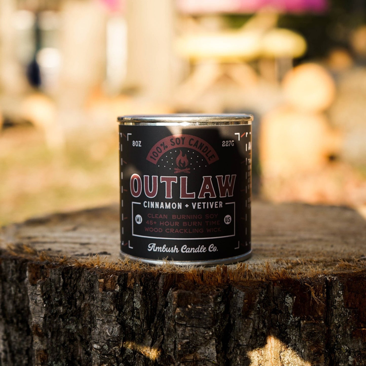 A can of Outlaw soy candle with cinnamon and vetiver scents, placed on a wooden stump outdoors. The label on the can indicates the scent profile and brand.