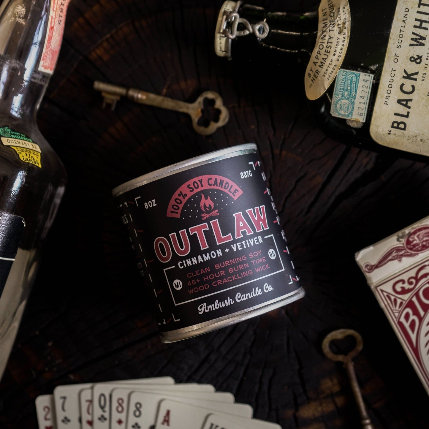 An 8oz soy candle with a label reading 'Outlaw Cinnamon & Vetiver', placed on a wooden surface surrounded by a key, a pack of cards, and some other items.