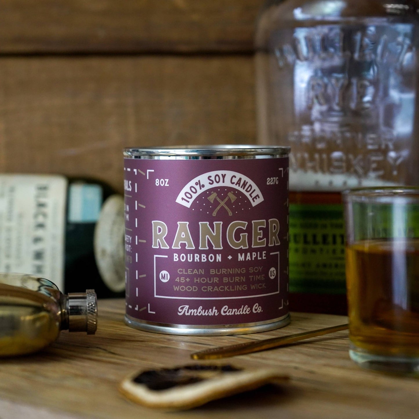 An 8oz soy candle labeled 'Ranger Bourbon Maple' with a maple leaf symbol, placed on a wooden surface next to a glass bottle and a metal container.