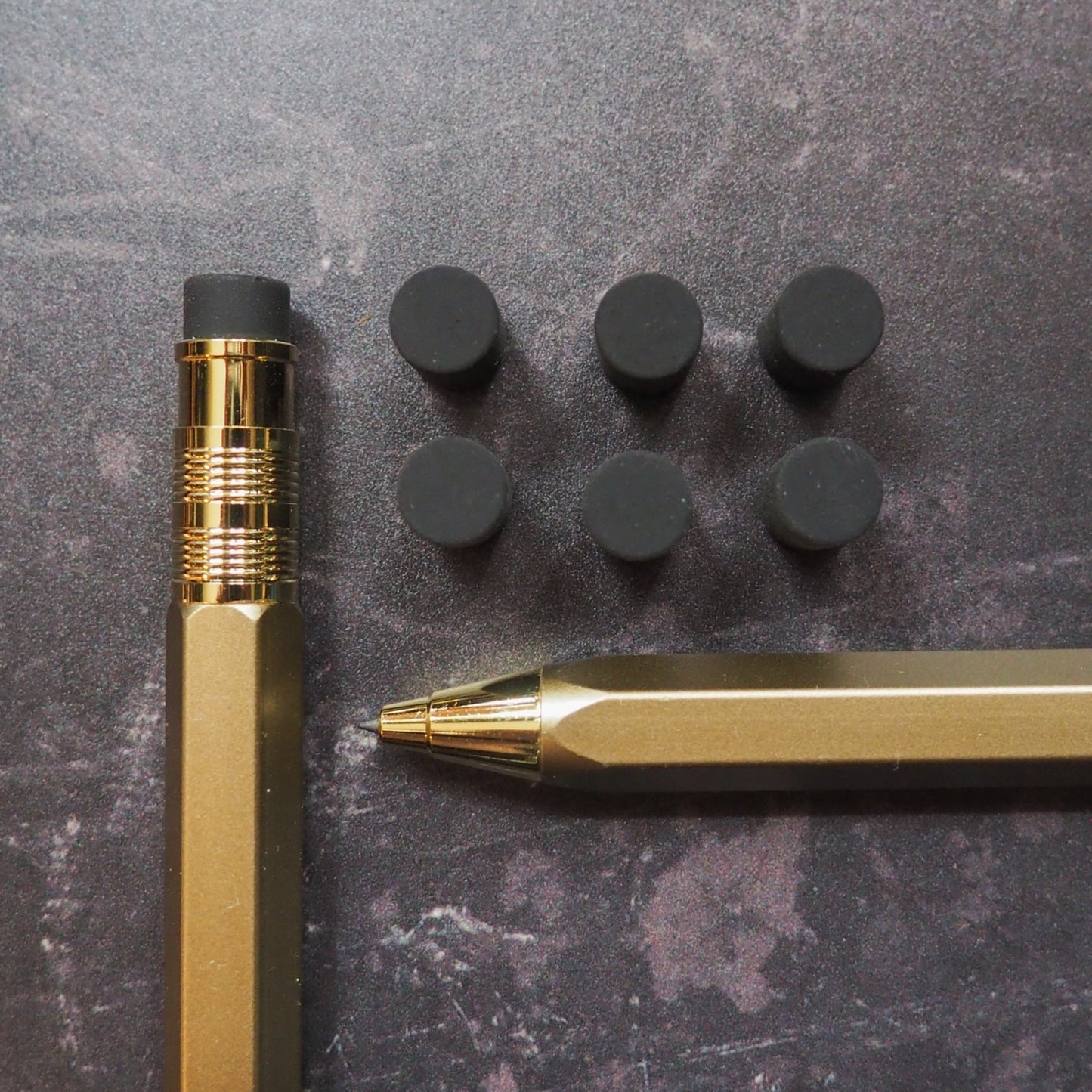 Gold pen and mechanical pencil with black erasers on a dark surface