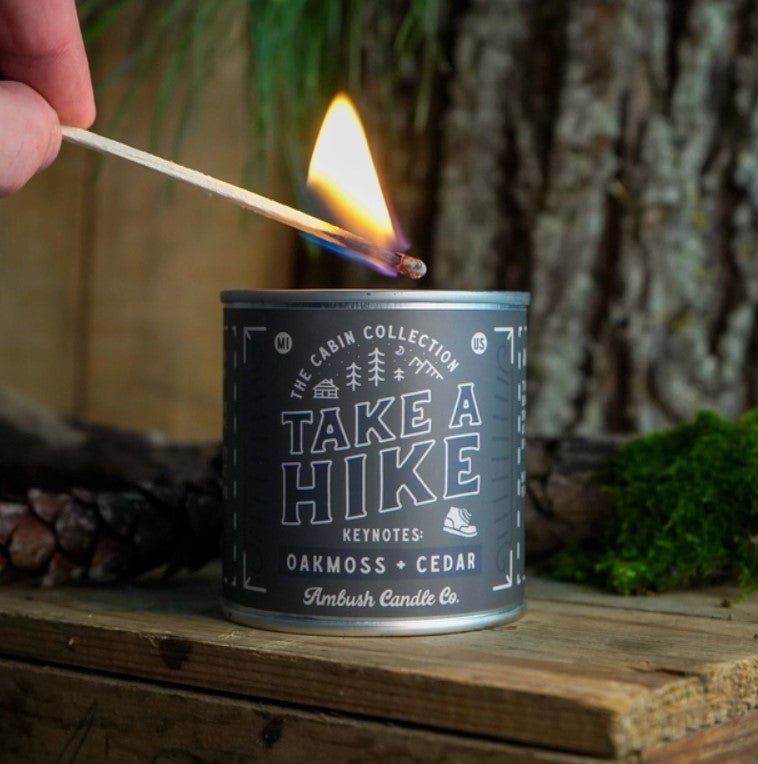 A candle with a label 'Take A Hike | Oakmoss + Cedar' is being lit with a match, placed on a wooden surface with a tree in the background.