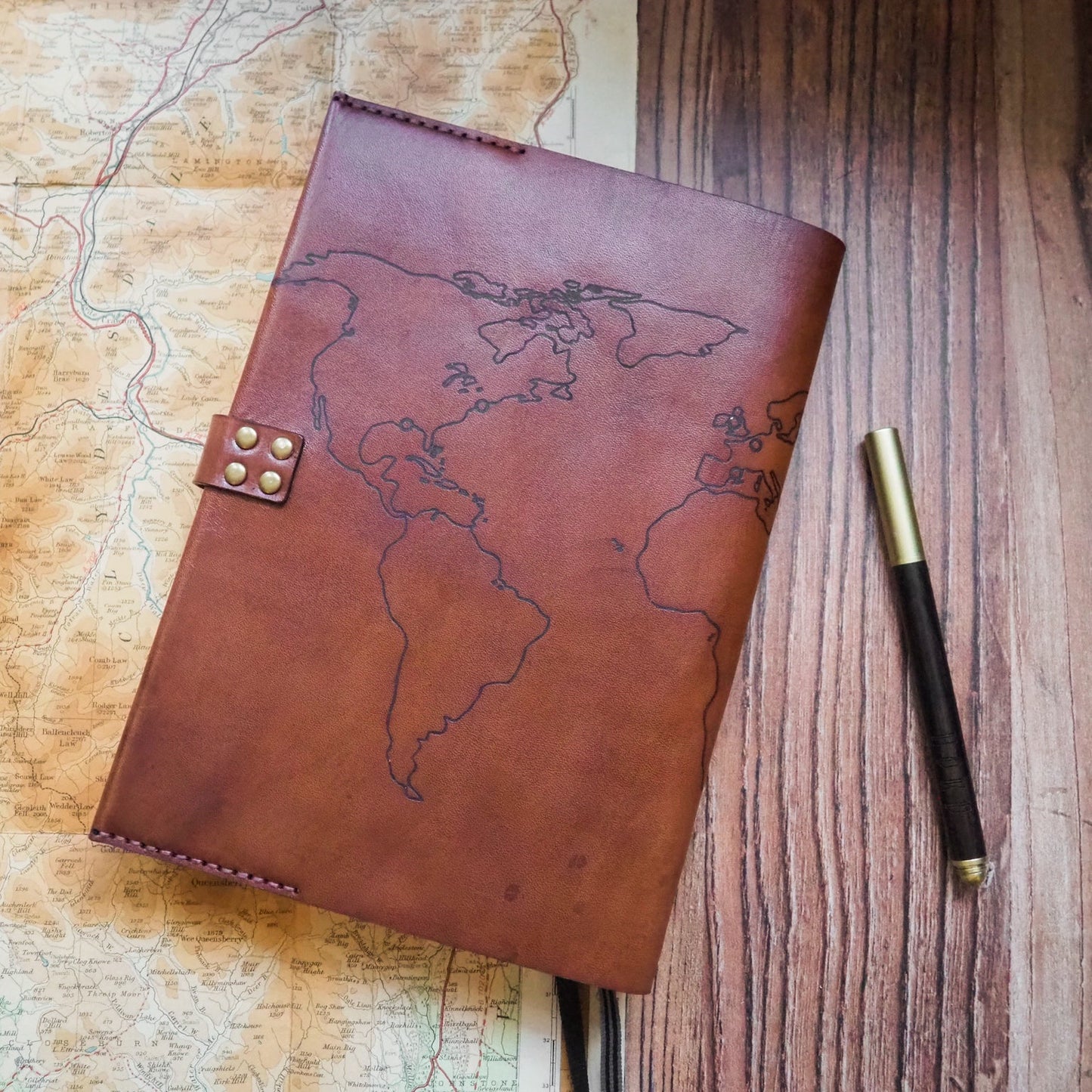 Brown leather notebook with world map design on a wooden surface with a pen beside it.
