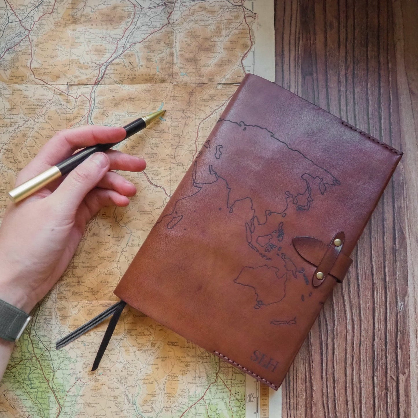 Brown leather notebook with world map design on a wooden surface with a hand holding a pen.