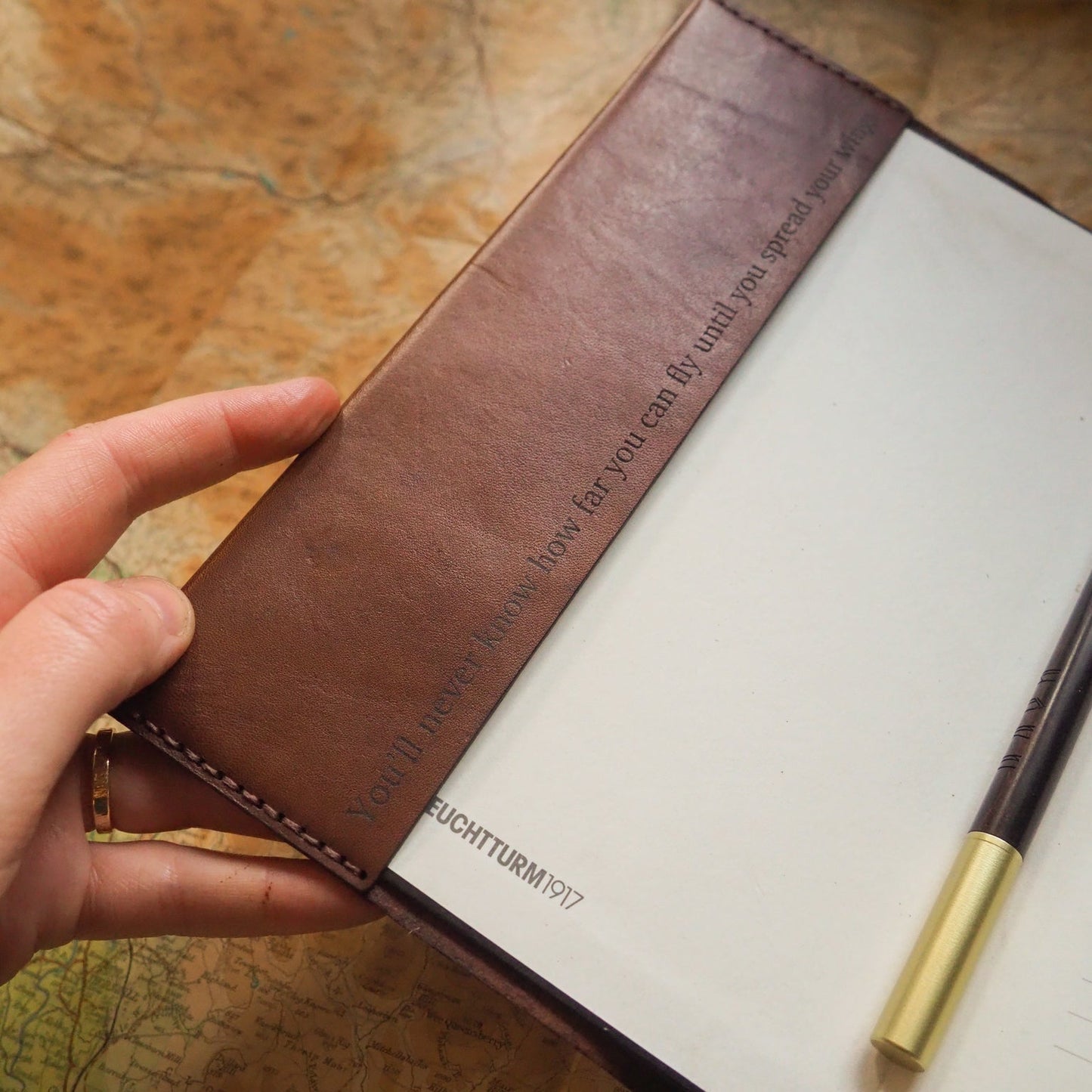 Hand holding a brown leather notebook  that's engraved with a s secret message with a pen on a map background