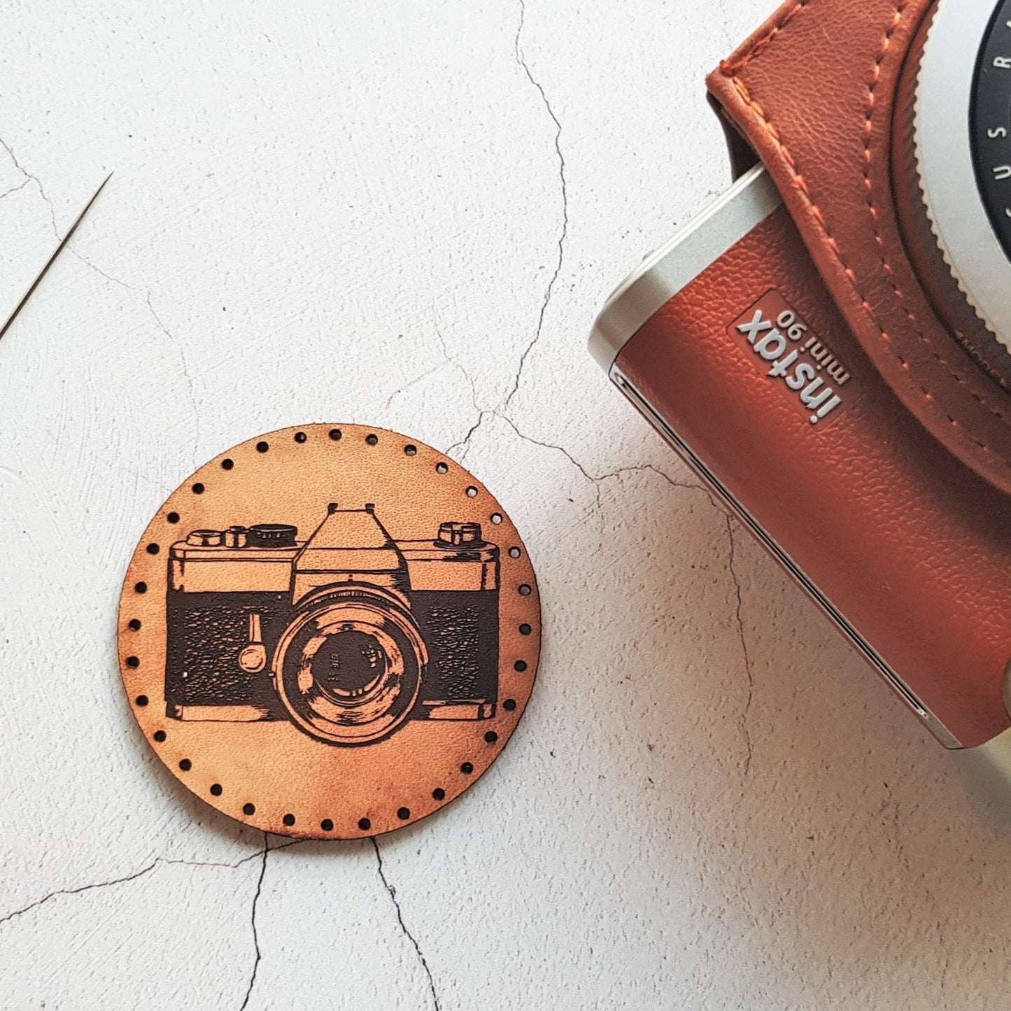 Circular leather patch in a tan patina engraved in black with the image of a vintage camera - pictured with a classic instax camera, a photographer patch from Hord.
