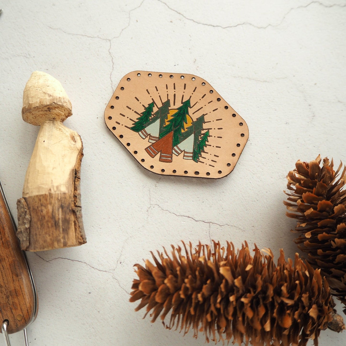 A luxurious leather patch, hand painted with green trees and brown tents with the yellow sun glowing in the background. The camping patch from Hord.