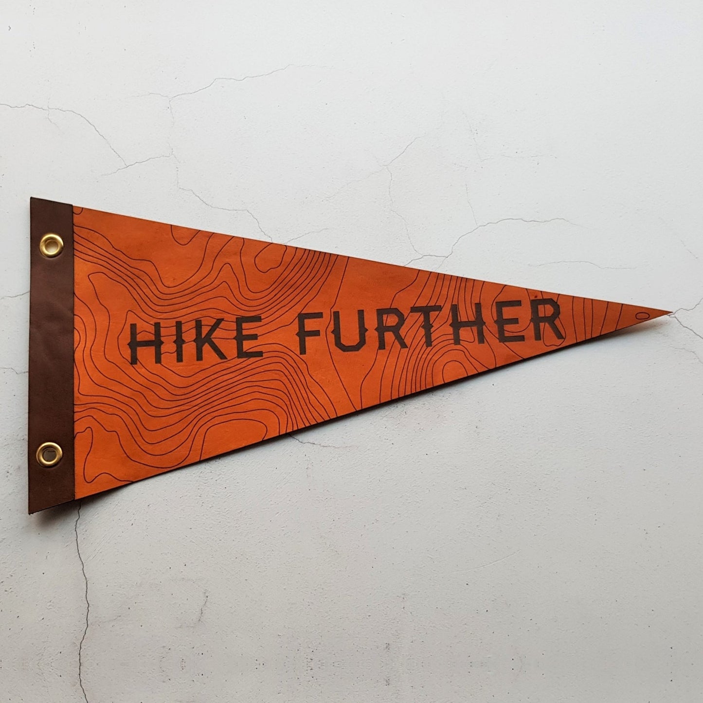 The Hike Further leather pennant, a hiking home decor by HORD is an inspiration piece of wall art with topographic map lines and the words 'Hike Further' on an orange tan background with a brown boarder.