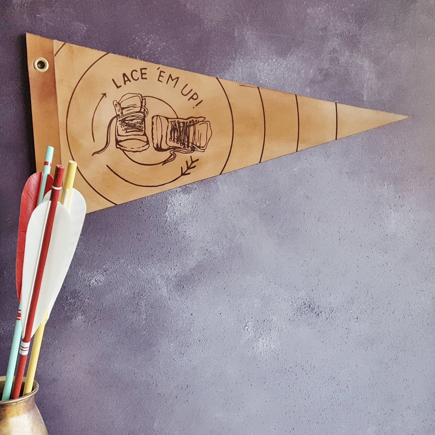 The Lace 'em up leather pennant, a camping home decor by HORD shows a pair of hiking boots on a light brown background, surrounded by a circular arrow motif and the words 'Lace 'Em Up!'
