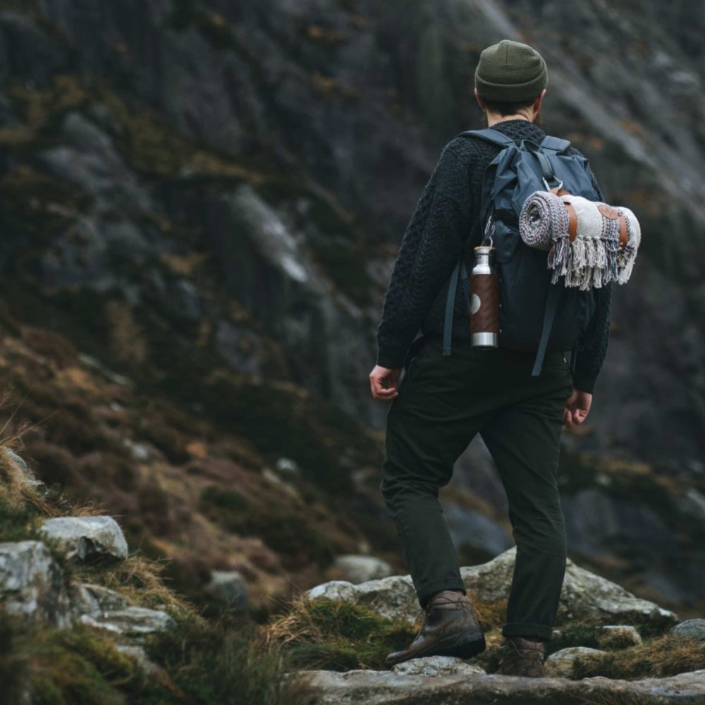 A man is walking up a mountain in Wales with a backpack on  that carries a recycled wool camper blanket and adventure bottle for gym