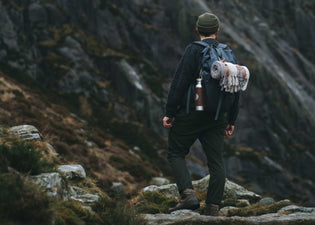  A man is walking up a mountain in Wales with a backpack on  that carries a recycled wool camper blanket and adventure bottle for gym