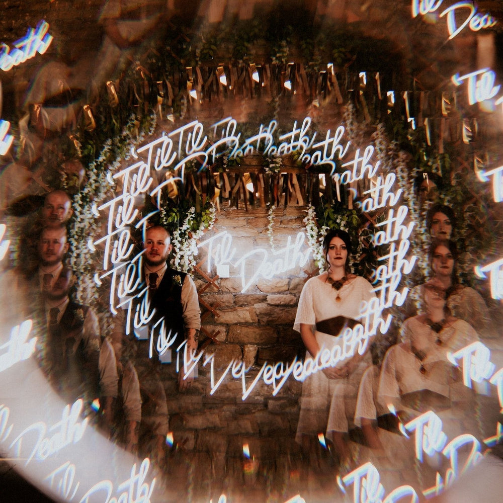 Jason and Gemma stands in the banquet barn for their wedding in Oakwell Hall, they stand in front of wisteria and a neon sign that says Til Death. The image is photographed to look like a kaleidoscope.