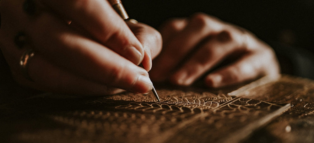 Gemma hand engraving some leather with a fern leaf pattern, using an awl. Moody lighting.