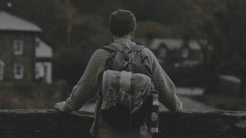 A hiker stands on a bridge in Wales with a backpack on his back, an insulated water bottle is and a recycled picnic blanket  is clipped to the bag