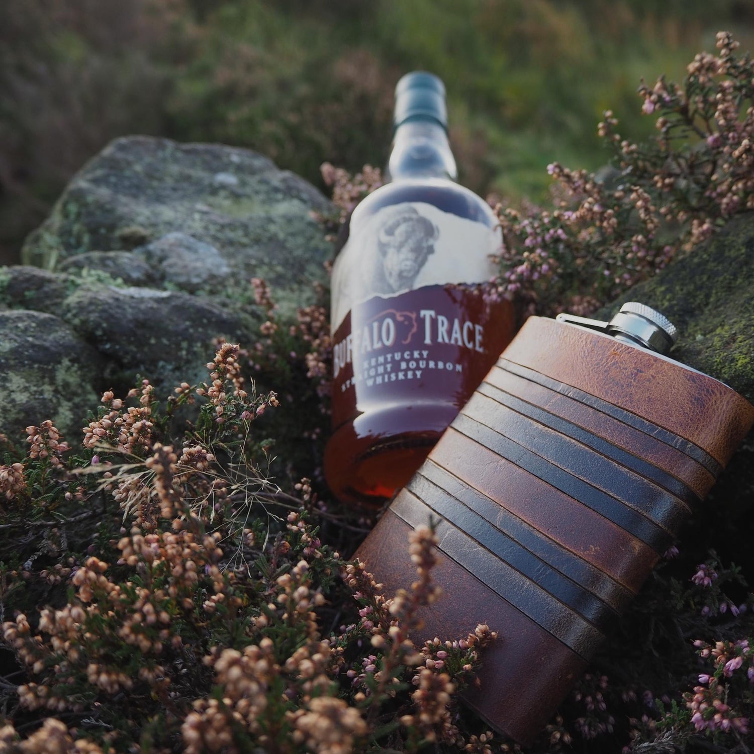 Our rust layers hip flask next to a bottle of whiskey, nestled in heather and stone on the moors.