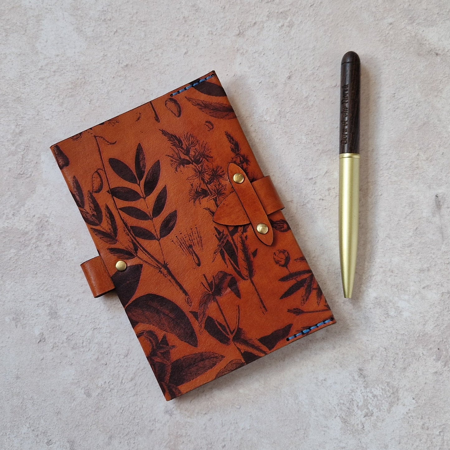 A leather journal with a botanical pattern in orange-brown and black, shown on a light textured surface next to a gold and black pen.