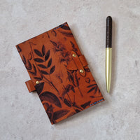 A leather journal with a botanical pattern in orange-brown and black, shown on a light textured surface next to a gold and black pen.