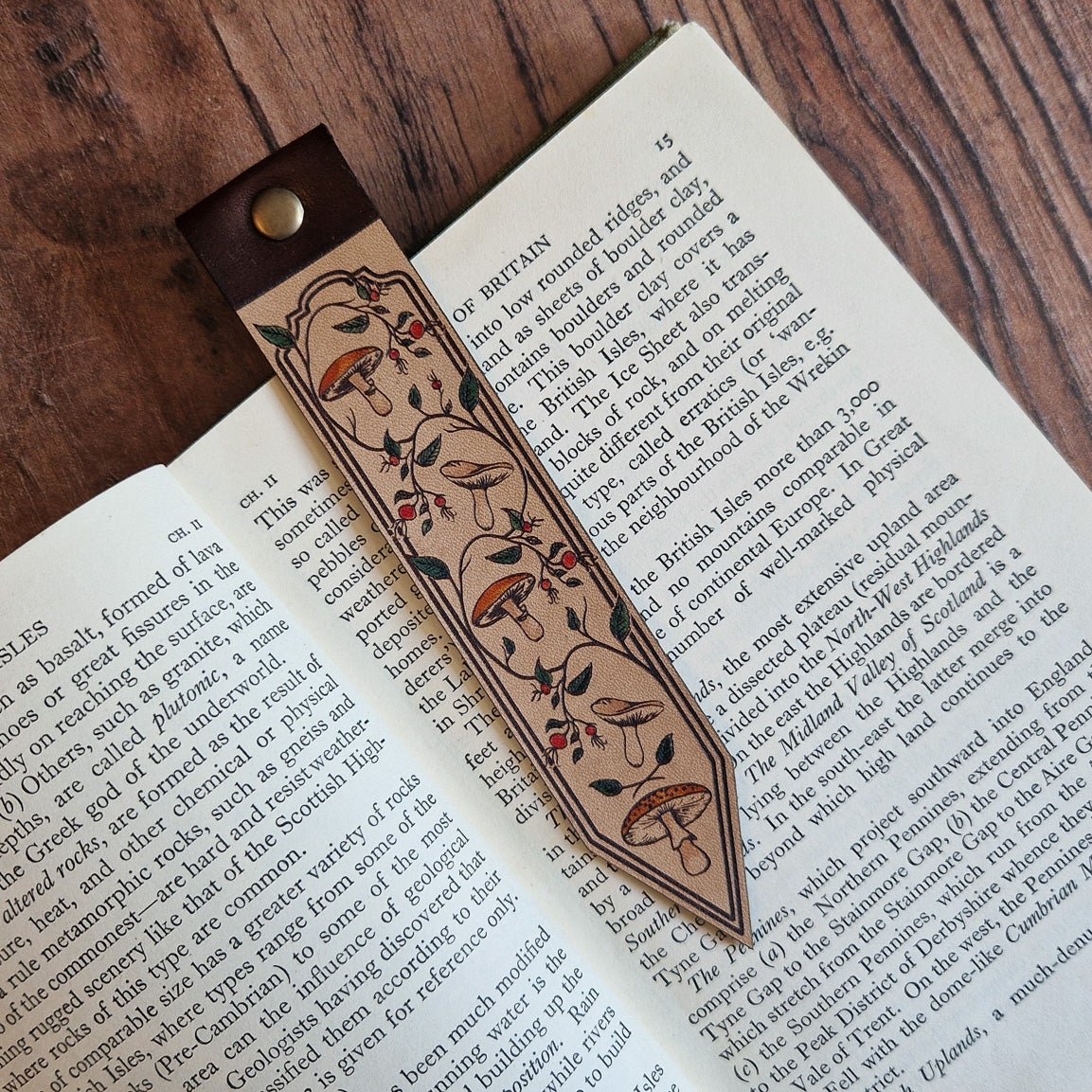 A decorative leather bookmark with floral engravings and a dark brown leather loop, resting on open book pages over a wooden tabletop.