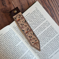 A decorative leather bookmark with floral engravings and a dark brown leather loop, resting on open book pages over a wooden tabletop.