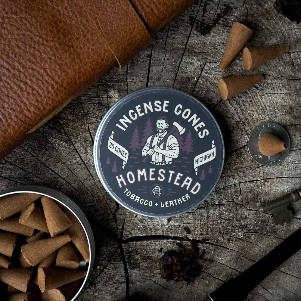 
                  
                    Incense cones container labeled 'Homestead' on a rustic wooden surface with incense cones and a pipe.
                  
                