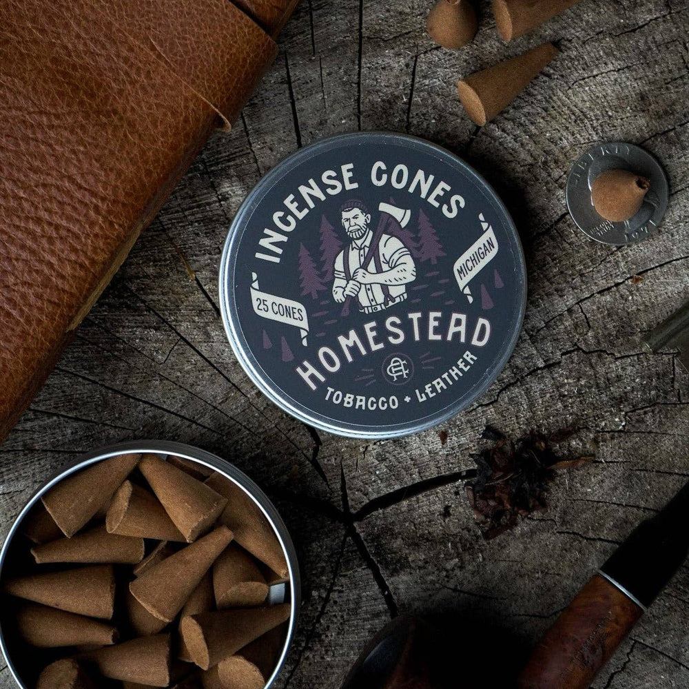 
                  
                    Homestead Incense Cones packaging on a wooden surface with incense cones and a pipe.
                  
                