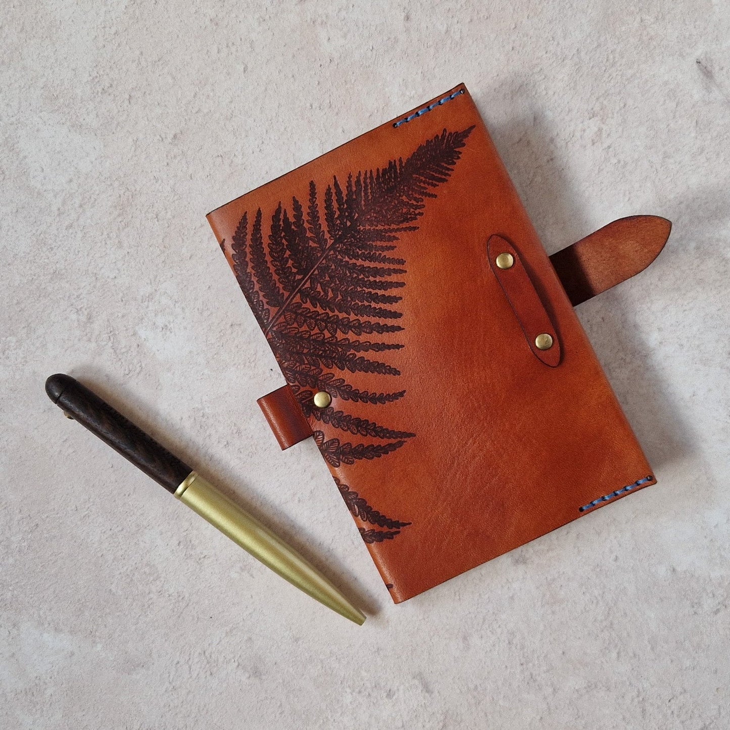 A brown leather journal with a dark brown fern leaf design on the cover, shown next to a gold and black pen on a textured light gray background.