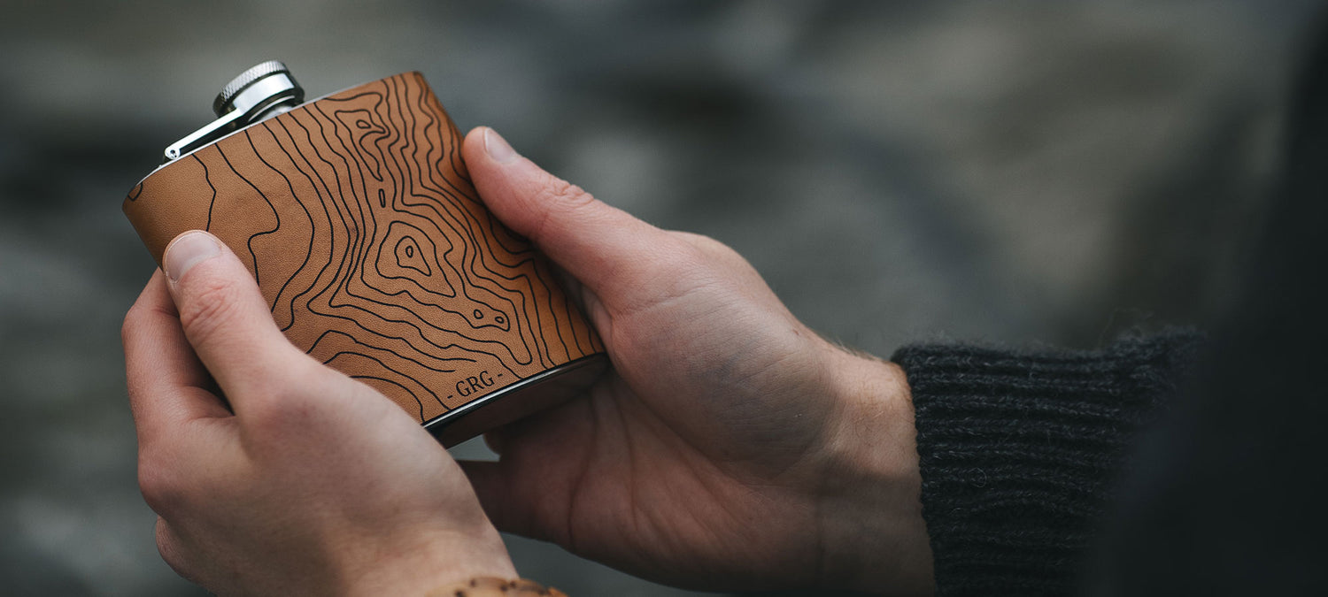 A person holds a leather clad hip flask engraved with the contour lines of their favourite place, by hord