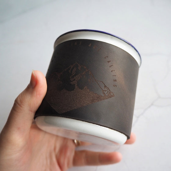 Our Mountains Are Calling illustration engraved on a leather wrapped engamel mug; an ideal gift for the camper in your life.