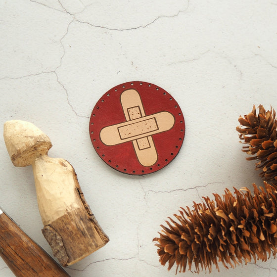 The First Aid patch by HORD is a circular leather patch featuring two crossed plasters on a hand painted red background, mirroring the universal first aid cross