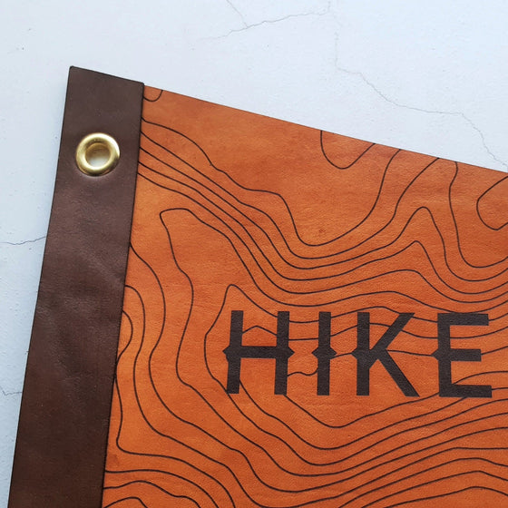 A close up of the Hike Further leather pennant by HORD, showing the texture of the black engrave on the hand dyed tan leather. Gold eyelets allow you to hang this flag style pennant up in the perfect place. A hiking home decor by Hord.