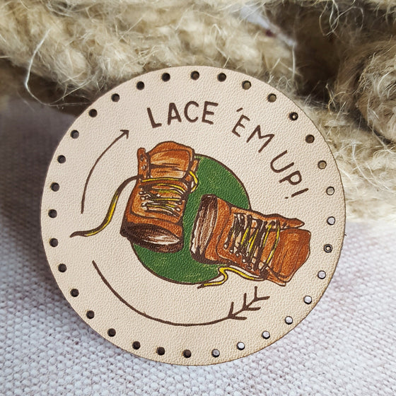 This circular leather outdoors patch is hand painted with a pair of brown hiking boots with yellow laces, surrounded by a green circle and text saying 'Lace em up!'