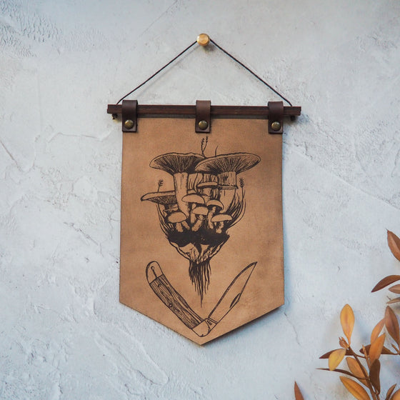 The Forager Banner by HORD is adorned with illustrations of a cluster of wild mushrooms and a survival knife. A great leather wall decoration piece.