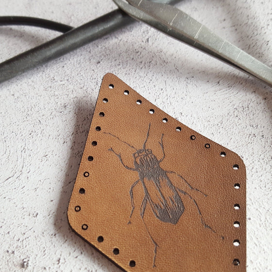 A close up of HORDs beetle patch, showing the texture of the grain of the leather and the depth of the black surface engraving. The beetle patch from Hord.