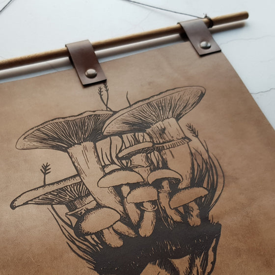 A close up of the Forager Banner, showing the details of the fungi illustration. Black engraving on antique brown leather with antique brass rivets and a wooden banner holder. A perfect leather wall decoration for your home.