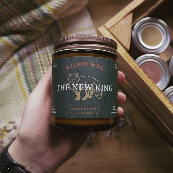 The New King Soy Candle, Hord. A hand holding an 8 ounce brown apothecary jar candle, A blanket and crate sits in the background