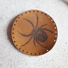 A close up photo of the HORD Spider patch from the entomology collection. A circular patch in dark brown leather with black engraving. The Spider Patch from Hord.
