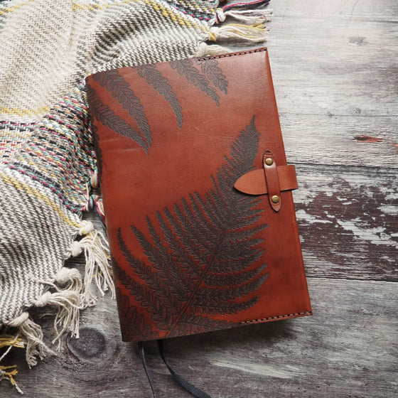 The Fern Leaf Leather Journal Cover by Hôrd in medium brown leather colour and light brown stitch colour. This leather book cover features a fern leaf illustration and has been tightened with a clasp.
