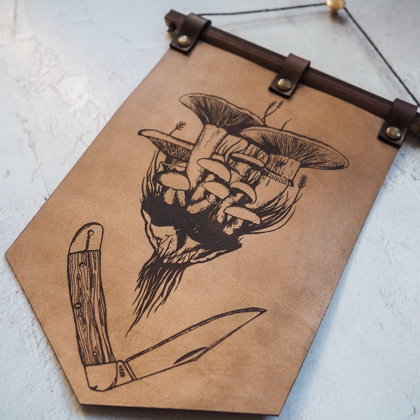 The forager banner, made of genuine full grain leather, wood and rivets. Depicting mushrooms and a foraging knife, a perfect leather wall decoration for your hallway.