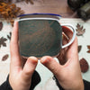 Hands holding an enamel mug wrapped with leather engraved with a mulberry leaf.